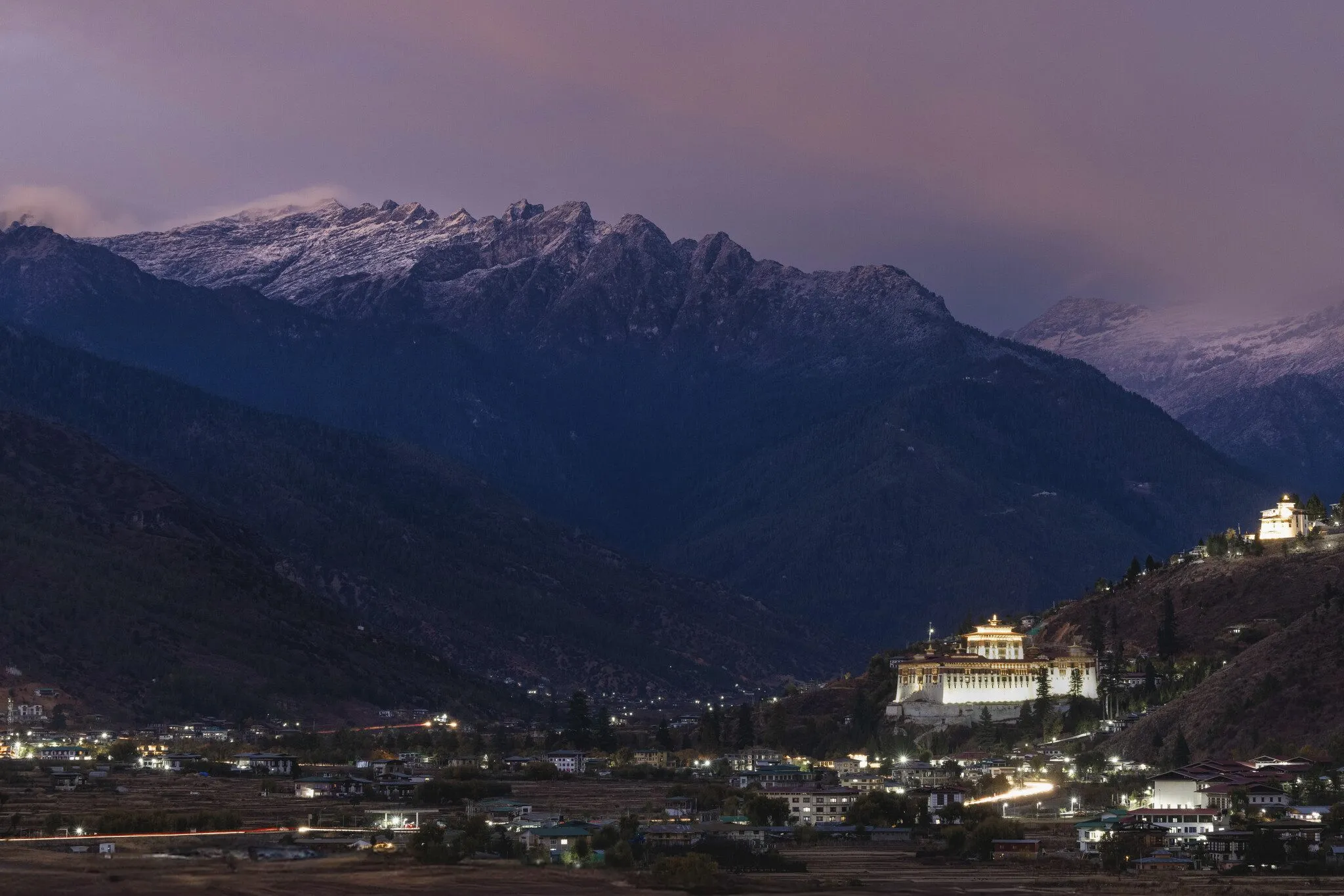 A floodlit dzong on a hillside above a lit valley town at dusk, with snow-dusted peaks behind