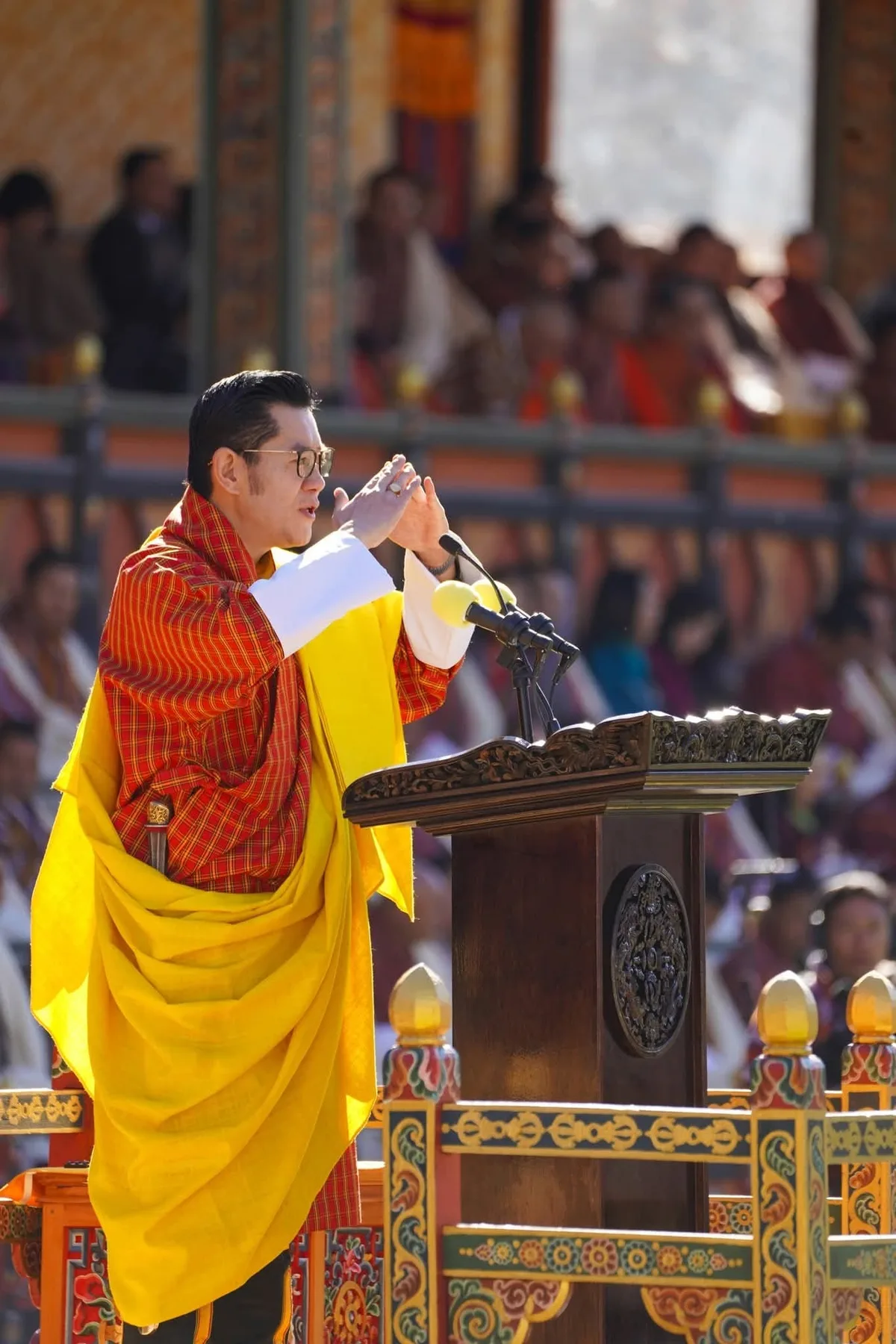 A speaker in a red gho and yellow scarf addresses a gathering from a carved wooden podium