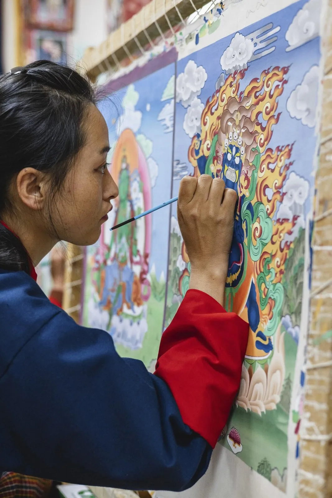 A young woman in a blue and red robe painting a detailed thangka of a deity among flames and clouds